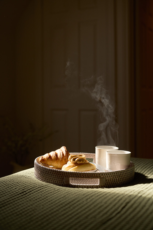a sunlit tray at the bottom of a bed with Croissants and 2 cups of coffee. the steam from the coffee is visible