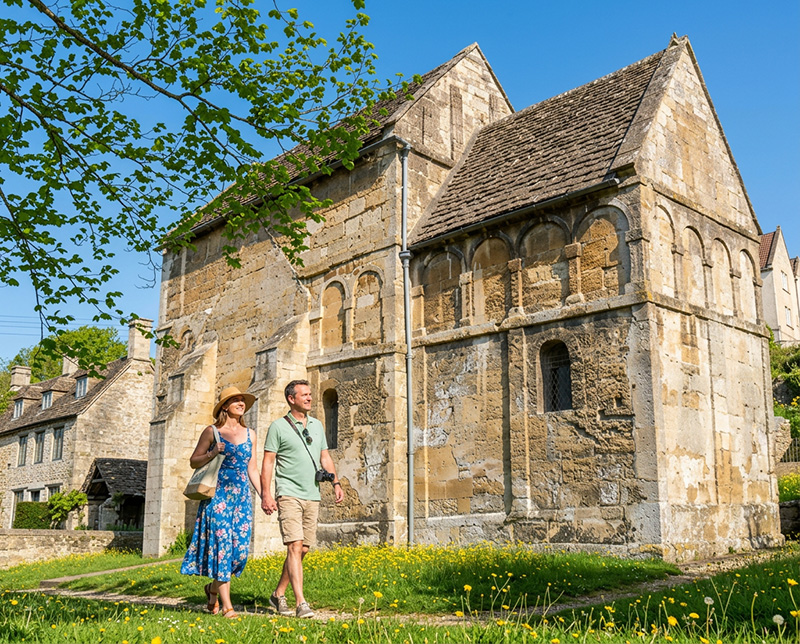 a couple walking passed St Laurence Church in Bradford-On-Avon on a Summers day